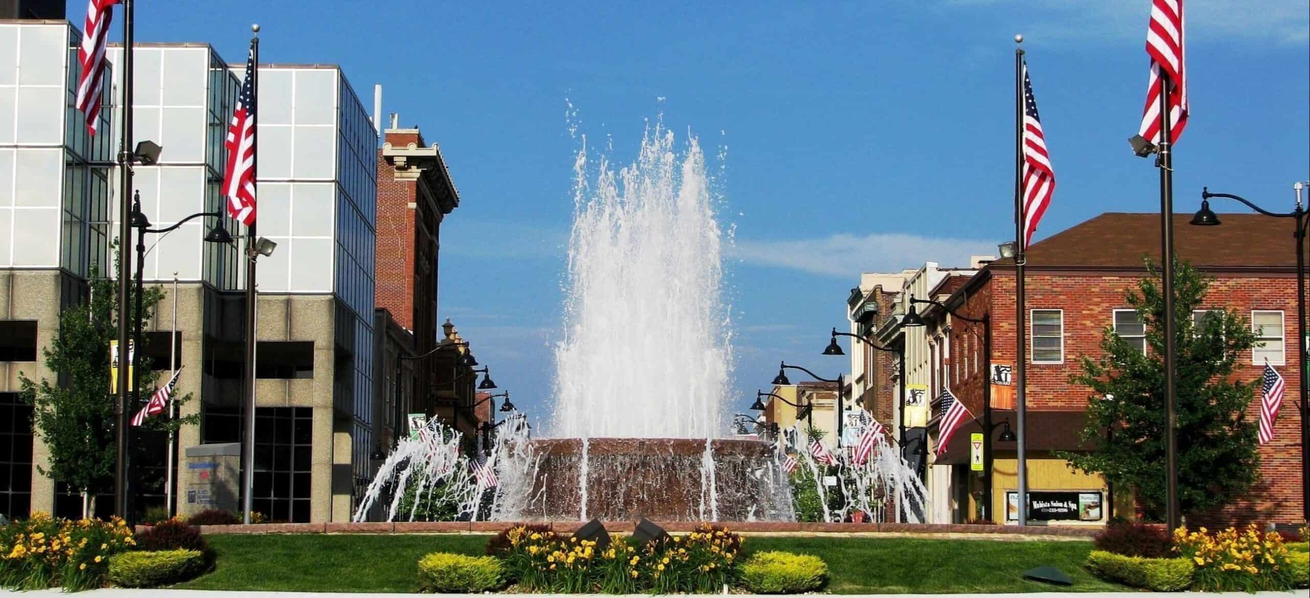 Fountain in a town square, surrounded by buildings and American flags
