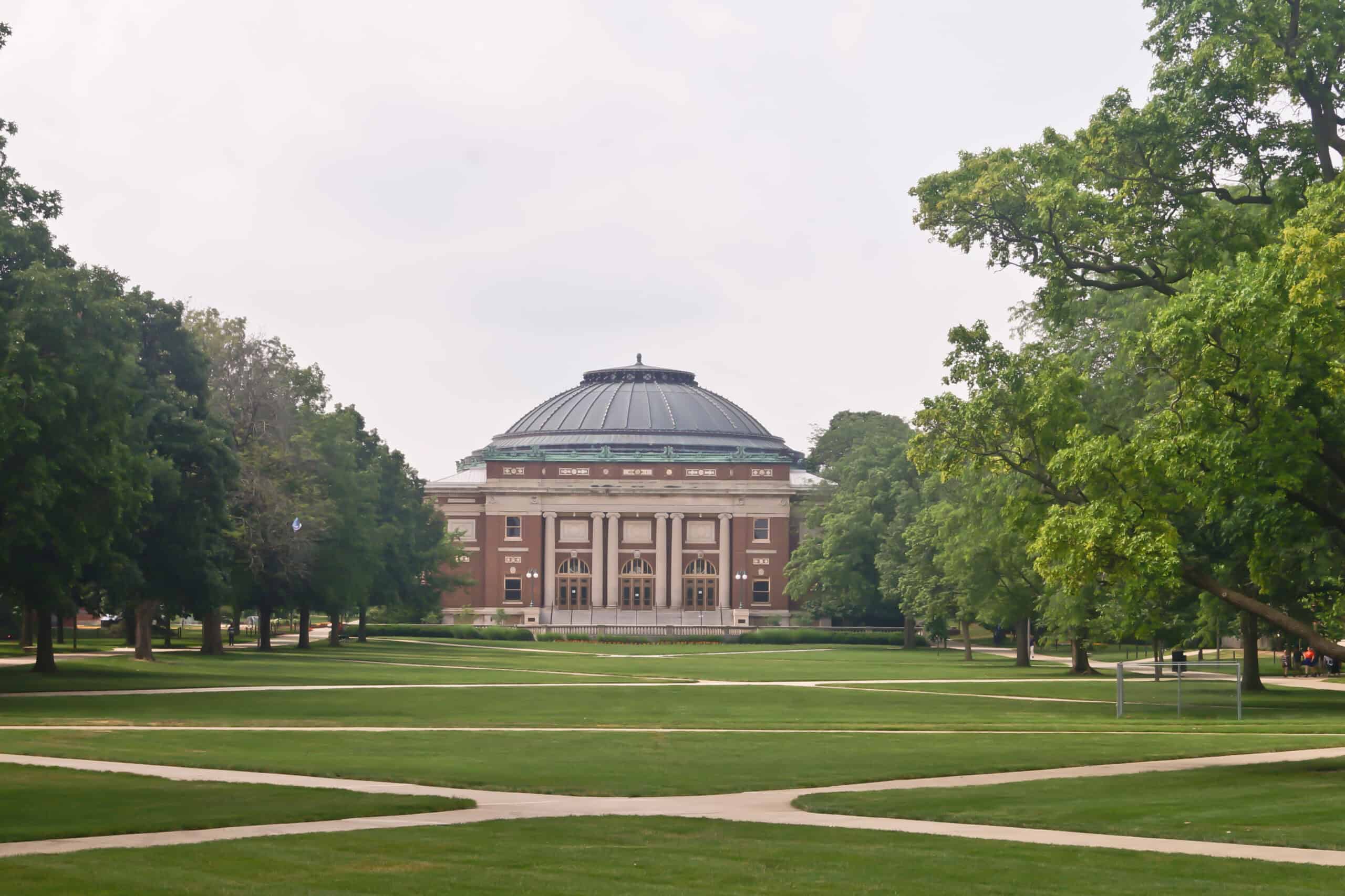 Image of a big lawn framed by trees with a tall brown building in the distance in the middle.