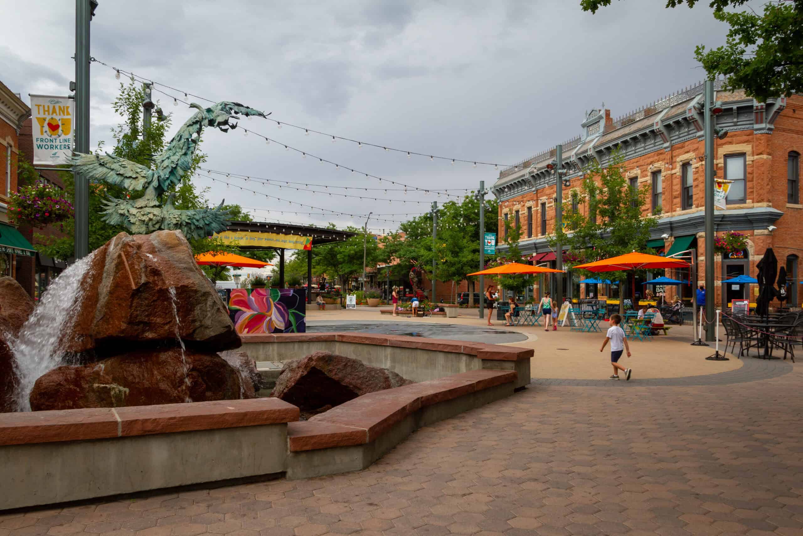 Close-up image of an area of downtown Fort Collins with an interesting building and some outside umbrellas with a centerpiece with brick benches on the left-hand side